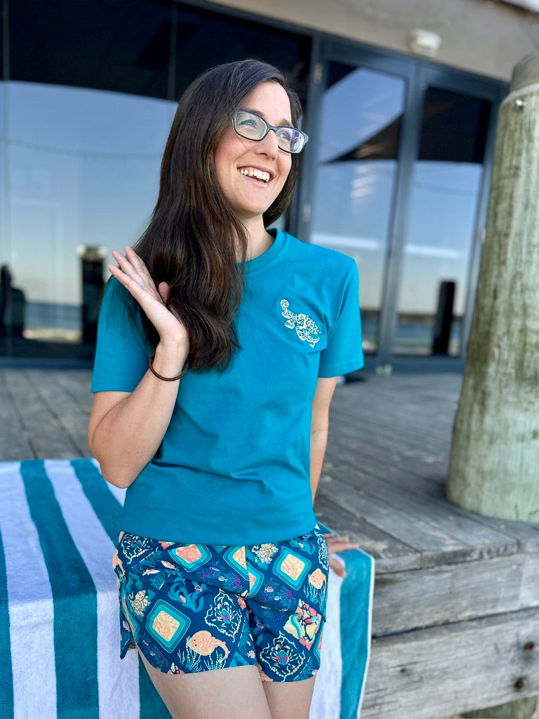 Woman wearing a blue t-shirt and patterned shorts sitting on a bench by a waterfront.