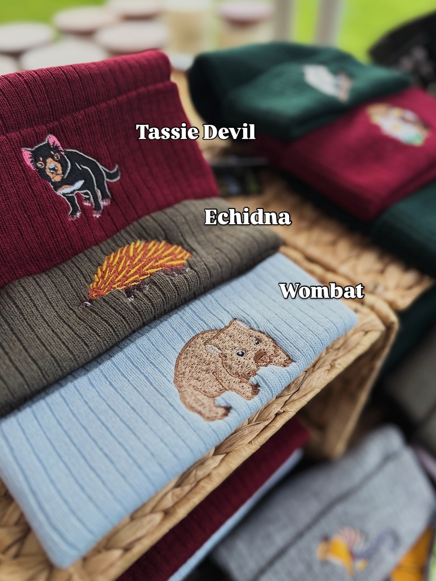 Row of beanies with embroidered animals labeled 'Tassie Devil', 'Echidna', and 'Wombat' on a woven box.