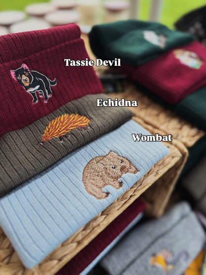 Row of beanies with embroidered animals labeled 'Tassie Devil', 'Echidna', and 'Wombat' on a woven box.