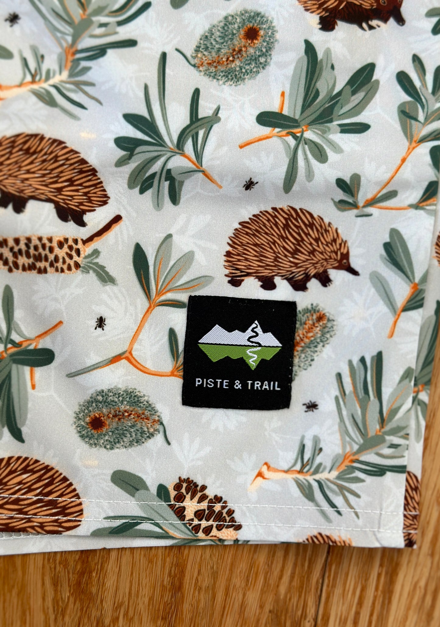 Shorts with echidna and banksia pattern and 'Piste & Trail' logo.