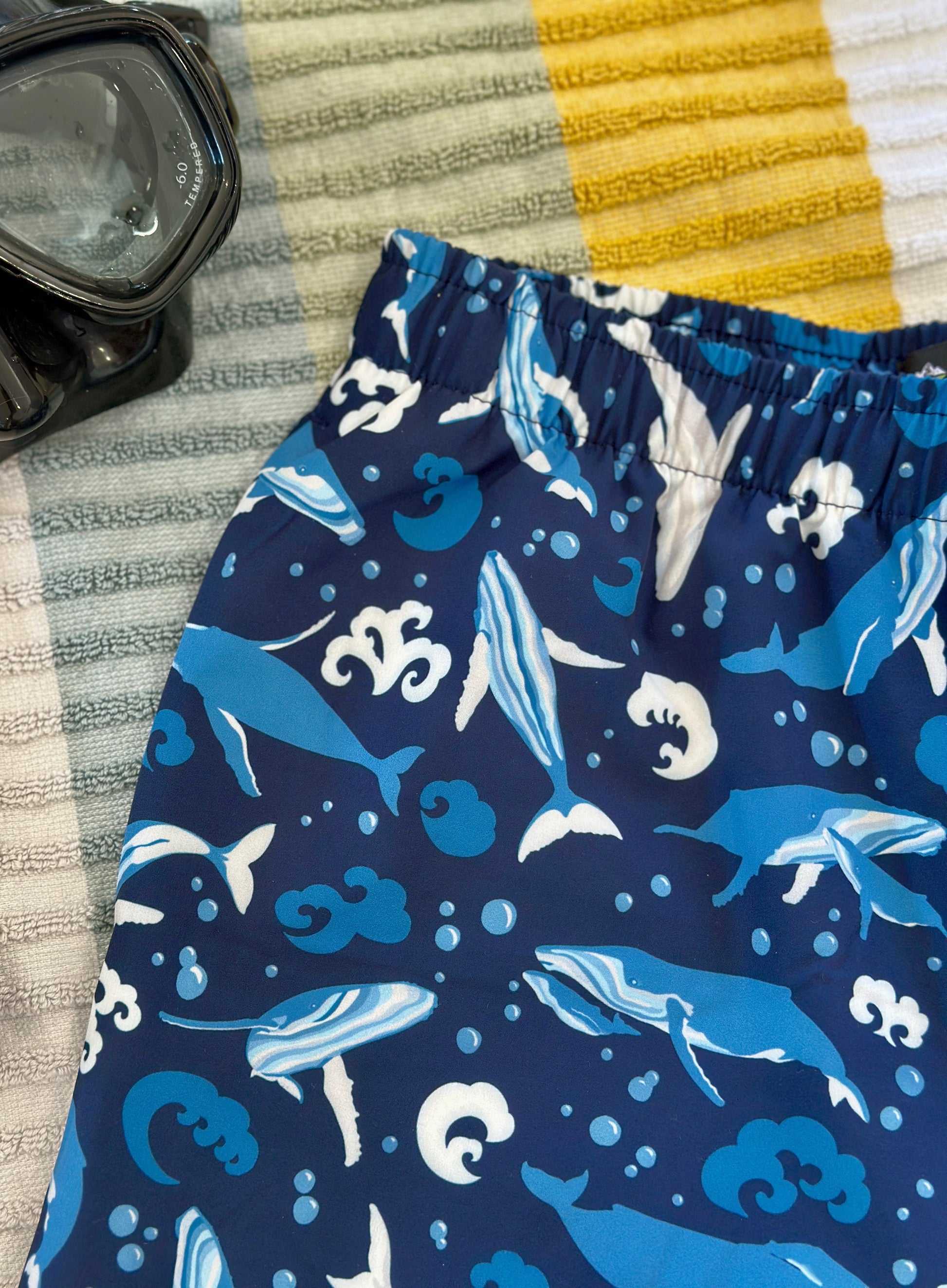 Navy swim shorts with whale pattern on a textured surface with goggles.