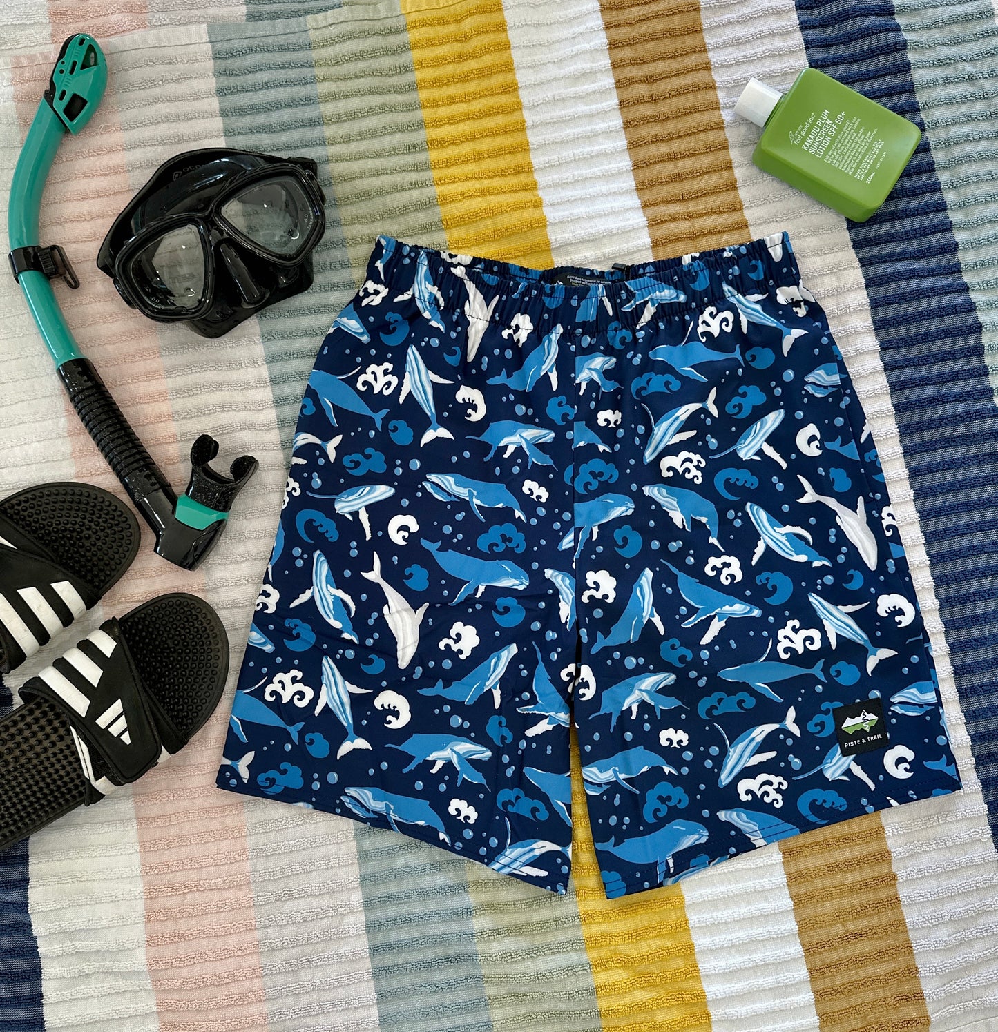 Navy blue swim shorts with whale pattern on a striped fabric background, surrounded by snorkeling gear.