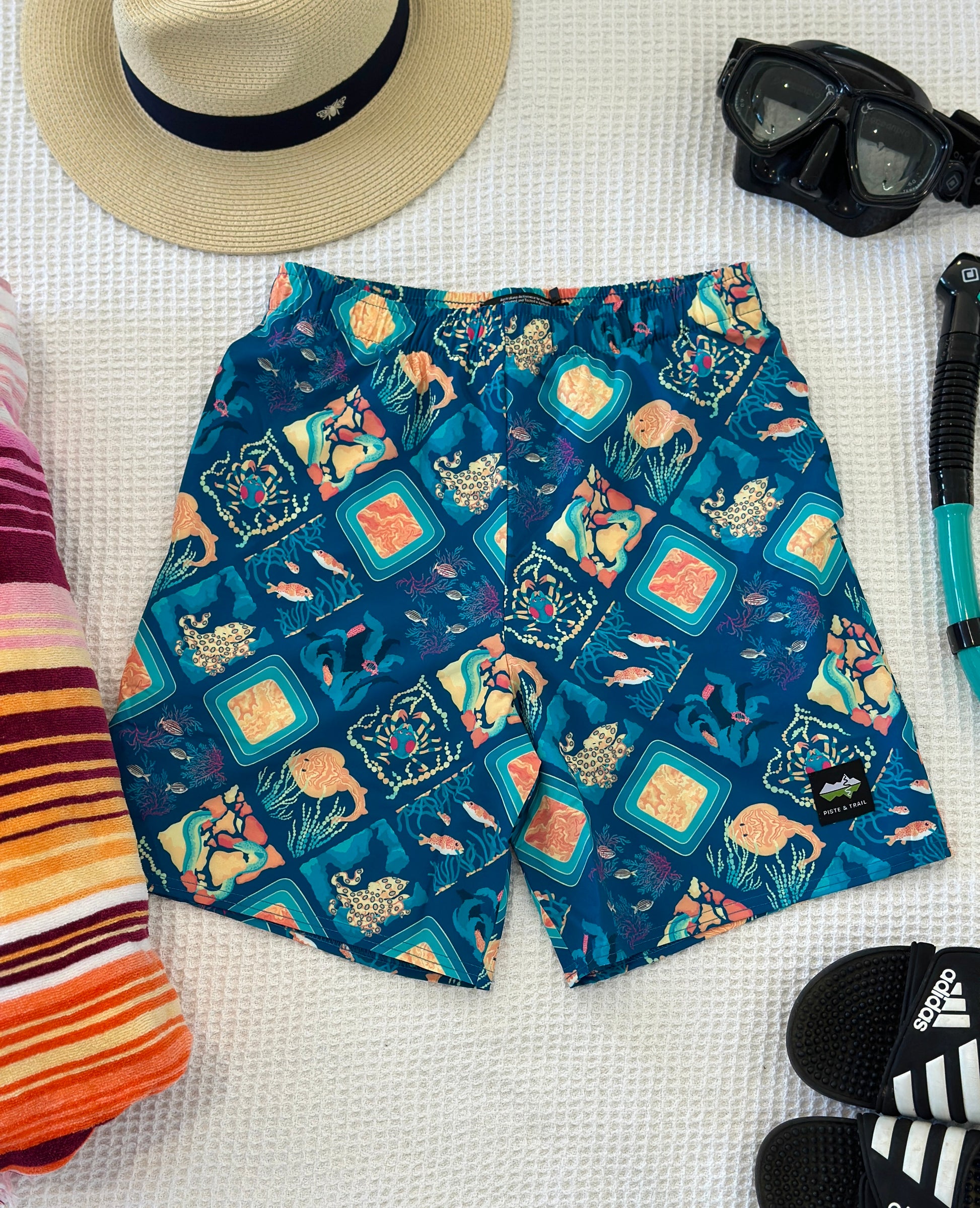 Colorful swim shorts with a marine life-pattern on a white surface, surrounded by a hat, goggles, and sandals.