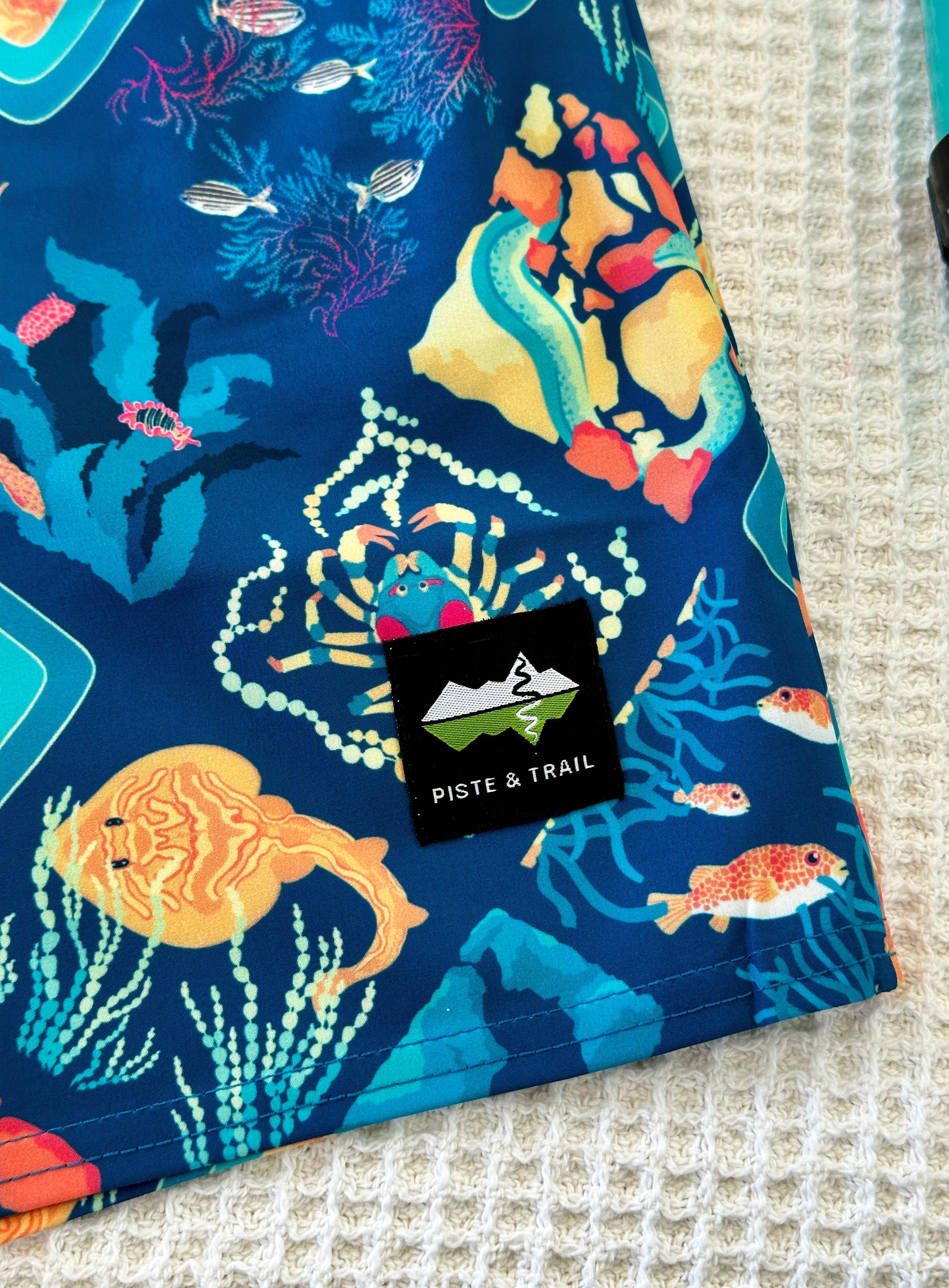 Blue fabric shorts with colorful ocean-themed design featuring fish, eels, and rays, with a Piste & Trail logo.