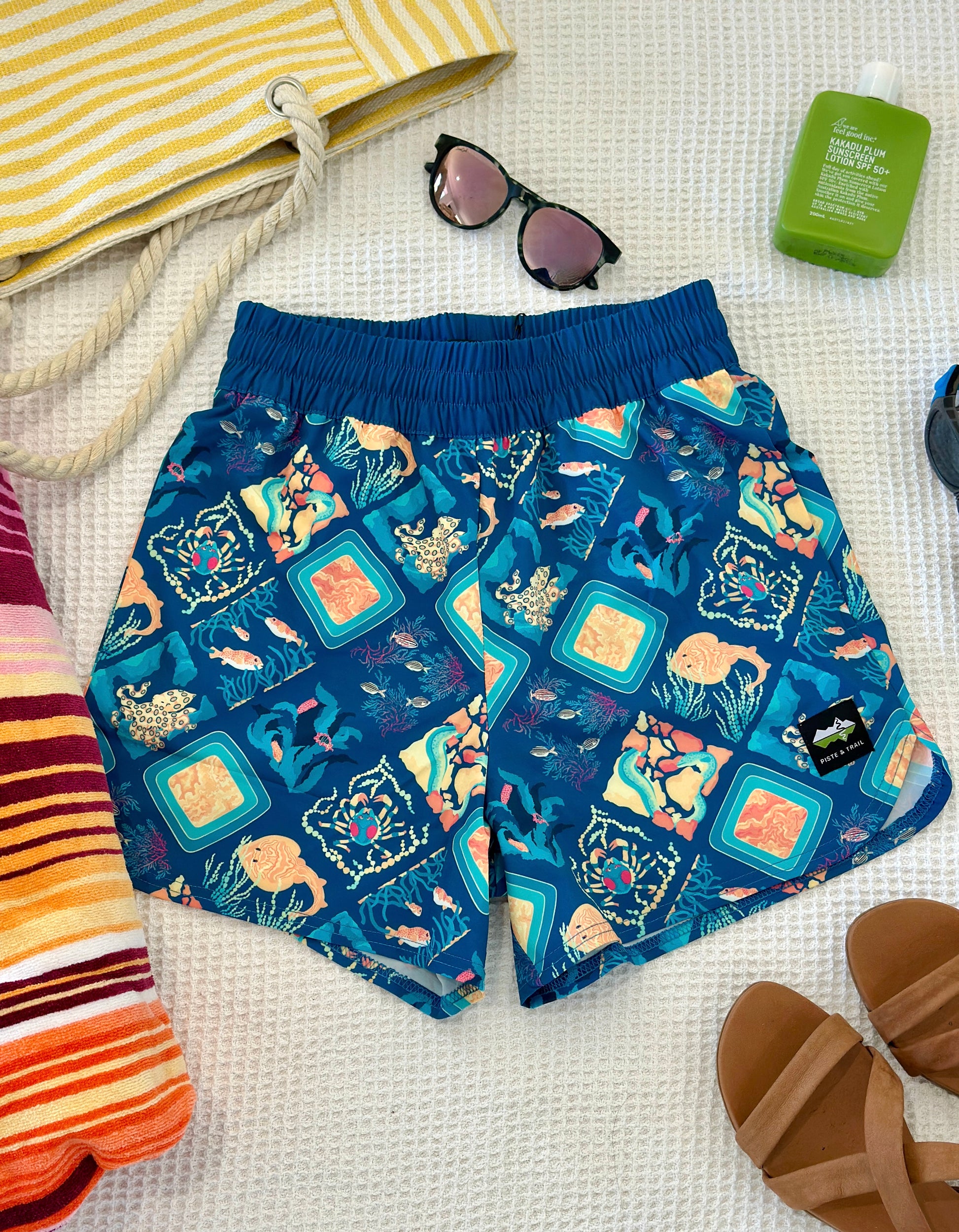Colorful marine life-patterned shorts on a textured surface with sunglasses, sandal, and towel.