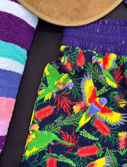 Colorful rainbow lorikeet-patterned shorts with a striped towel in the background