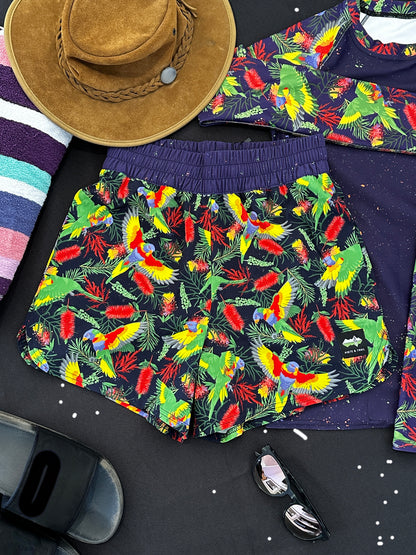 Colorful rainbow Lorikeet-patterned shorts with a hat and sunglasses on a dark surface