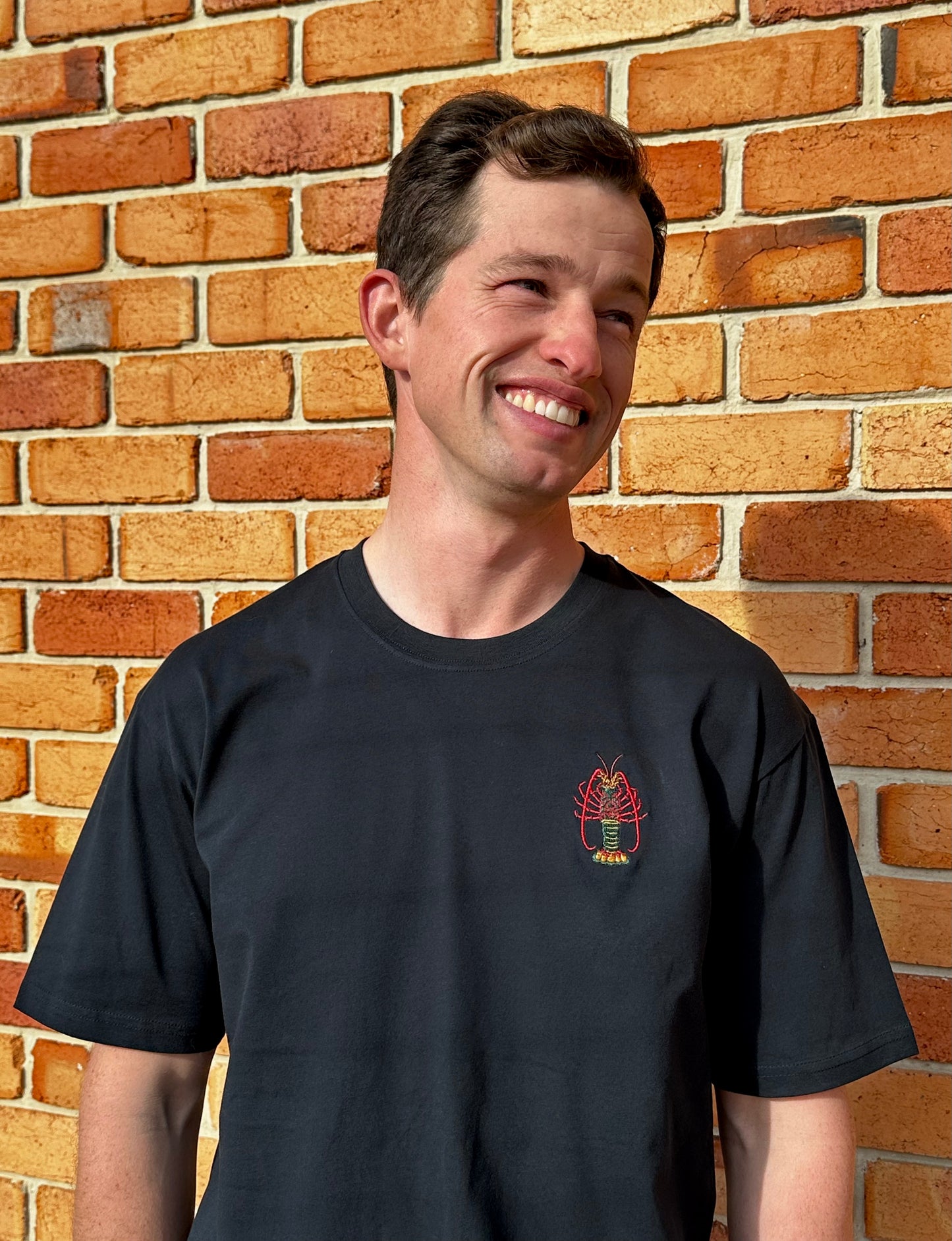 Man wearing a navy t-shirt with a lobster logo against a brick wall