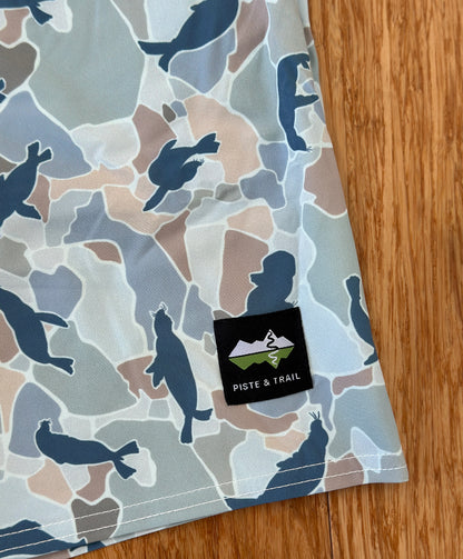 Camouflage-patterned board shorts with seal silhouettes with a Piste and Trail brand label on a wooden surface
