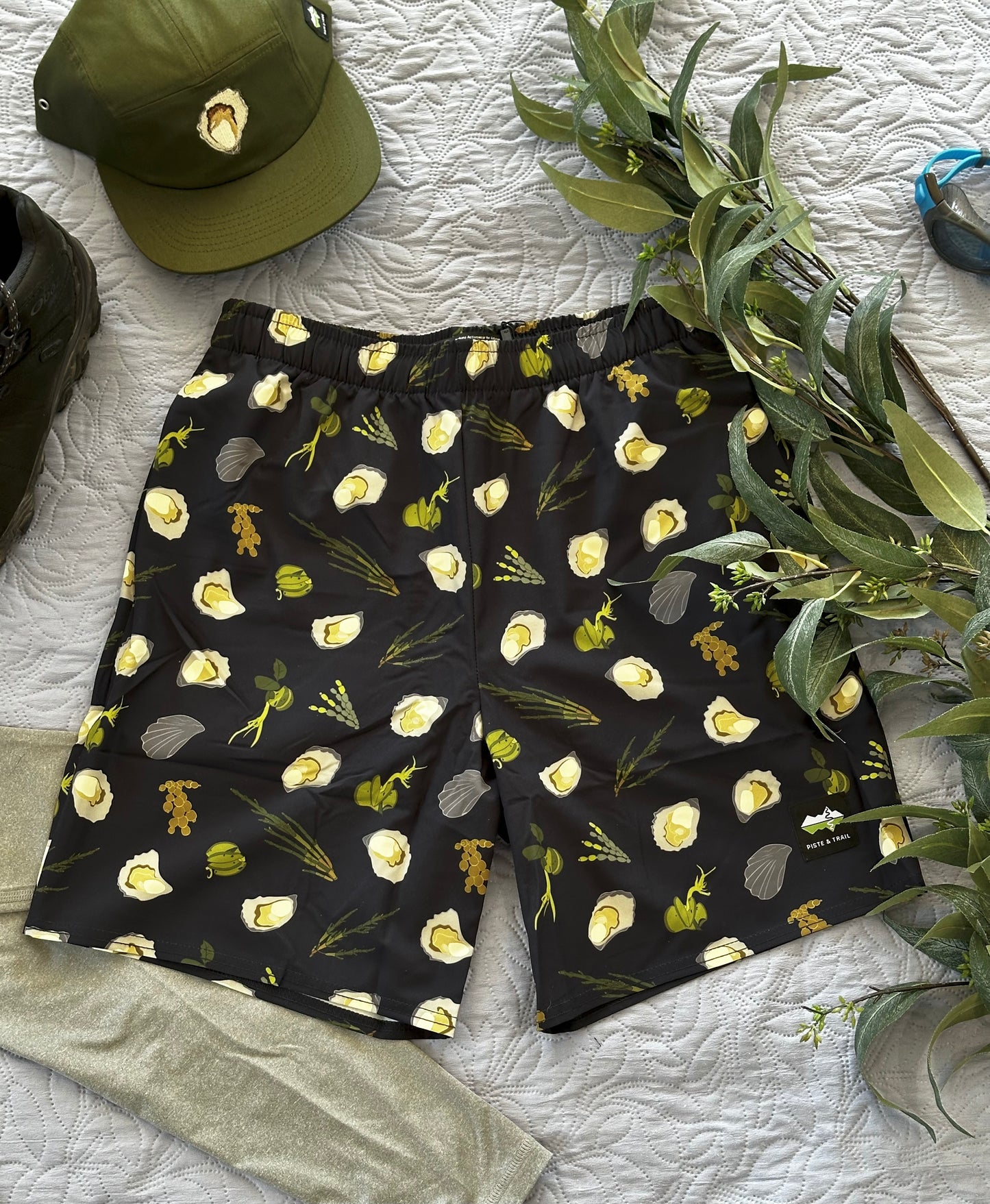 Dark Navy shorts with green seaweeds, and oysters pattern on a white textured surface with a green cap and plant.