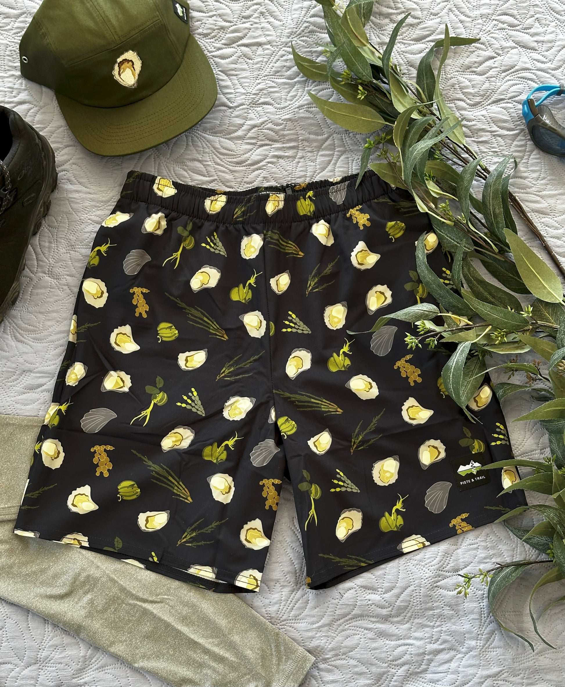 Dark Navy shorts with green seaweeds, and oysters pattern on a white textured surface with a green cap and plant.