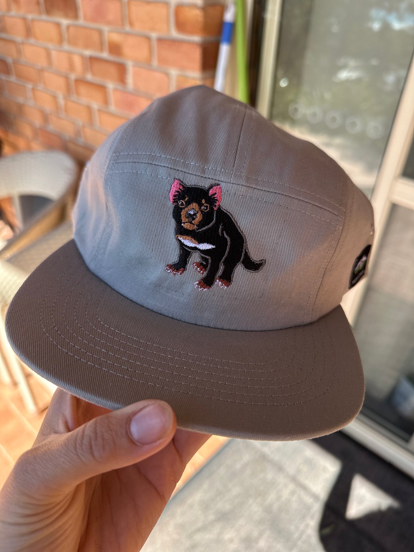 Gray cap with a black Tasmanian Devil embroidered design held by a hand outdoors.