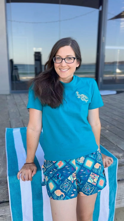 Woman wearing a blue shirt and patterned shorts sitting on a striped towel by the water.