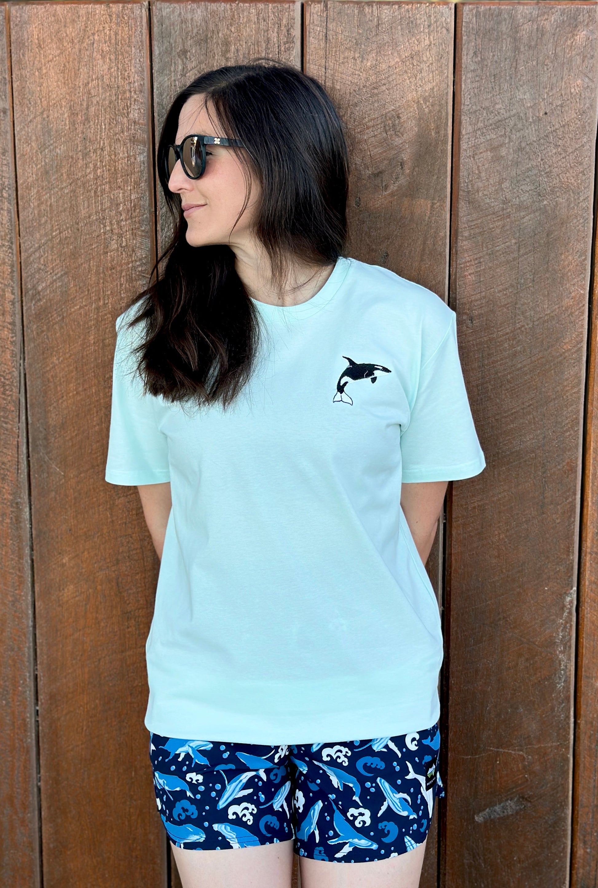 Woman wearing a light blue t-shirt with an orca design and blue shorts with humpback whale patterns, standing against a wooden fence.