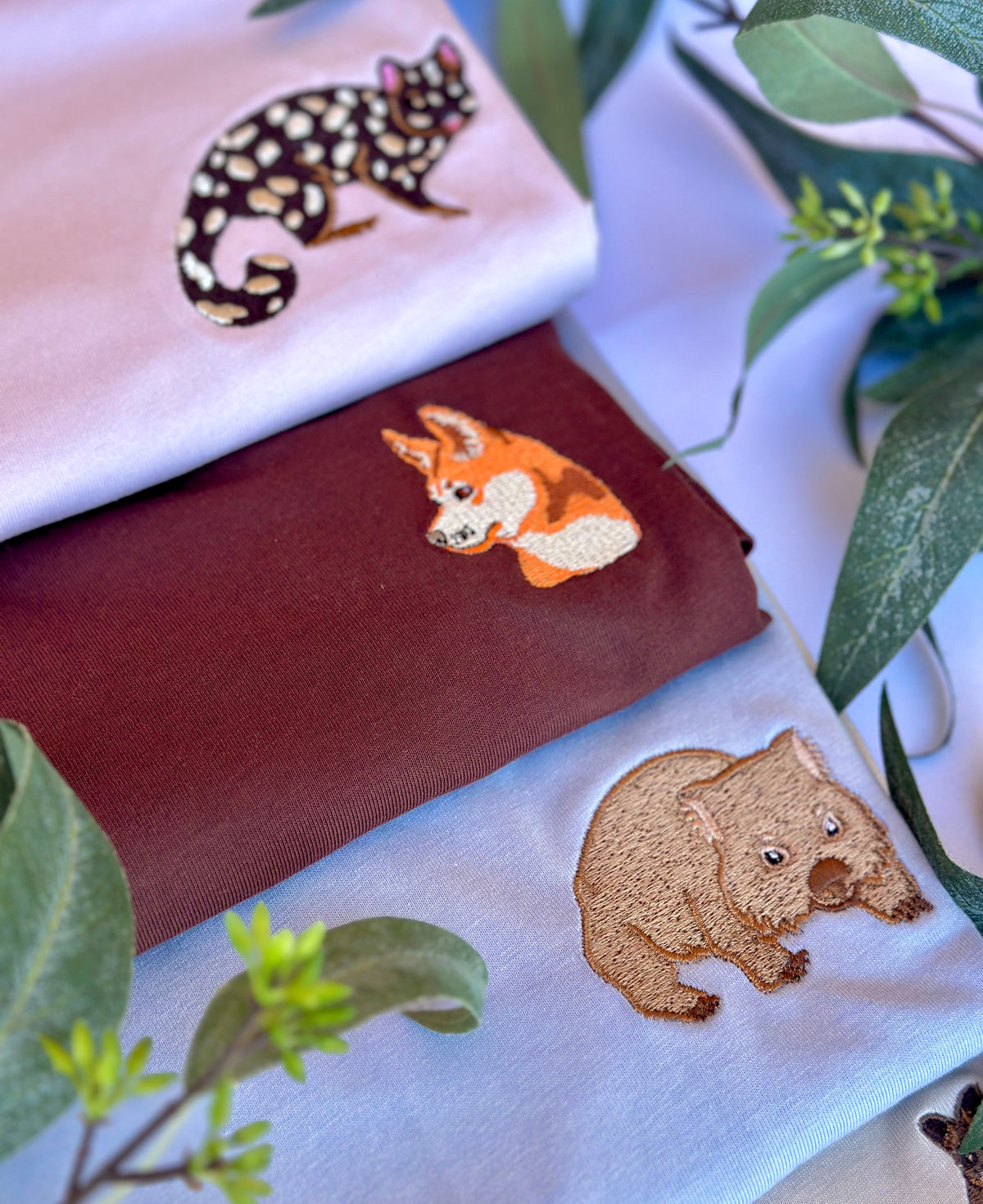 Three folded fabric items in pale pink, dark brown, and light blue, each embroidered with a different animal patch design (a spotted quoll, a dingo, and a wombat), styled with green leaves.