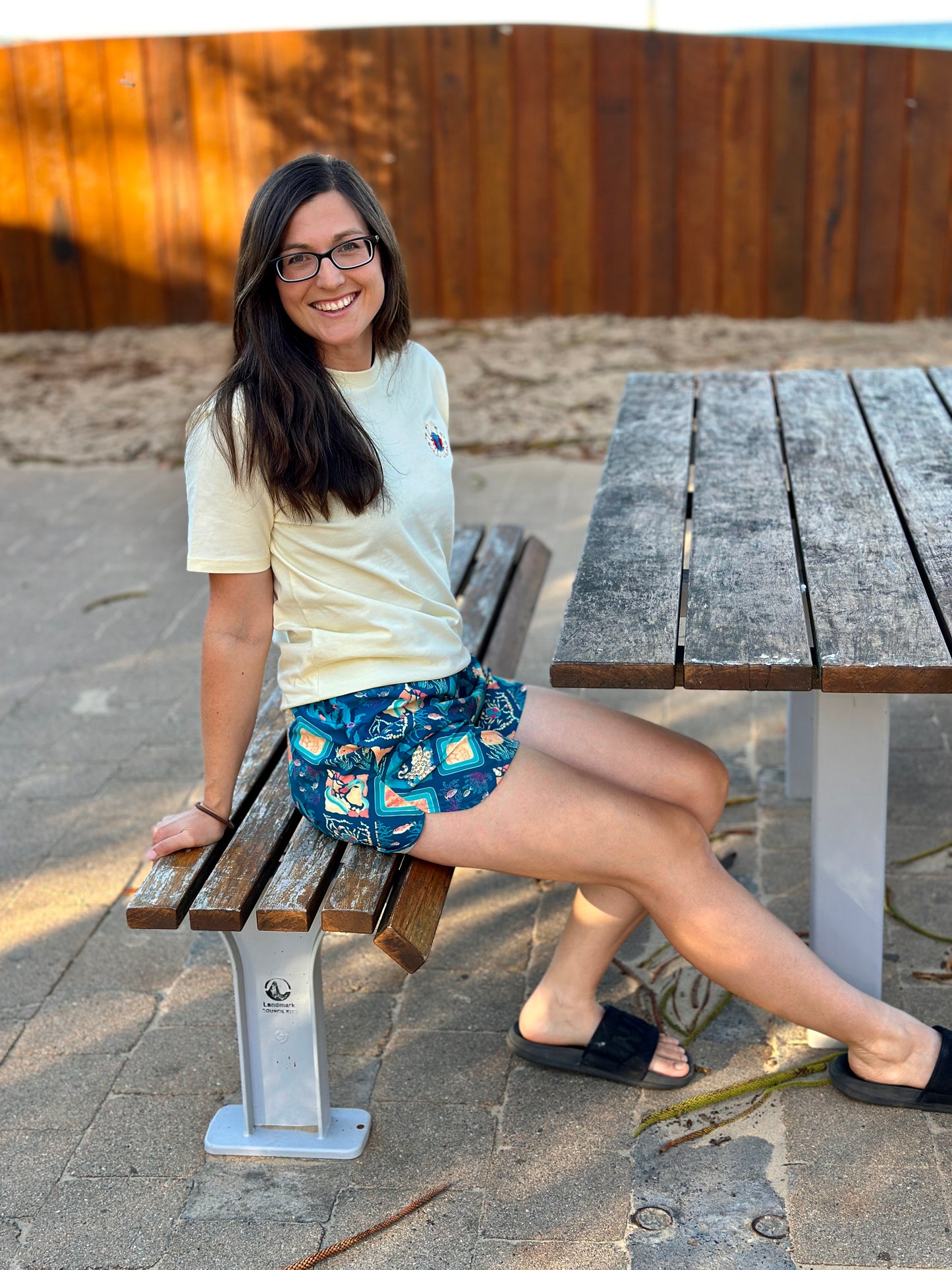 Woman sitting on a wooden bench at a park table, wearing a butter yellow shirt and patterned shorts.