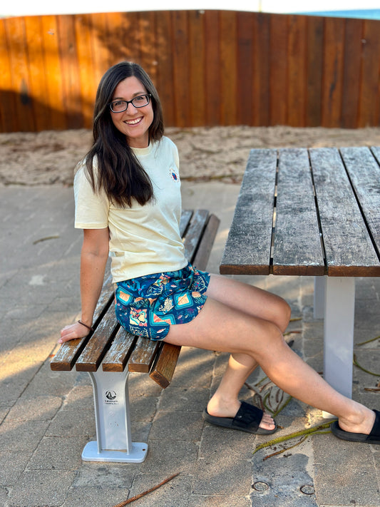 Woman sitting on a wooden bench at a park table, wearing a butter yellow shirt and patterned shorts.
