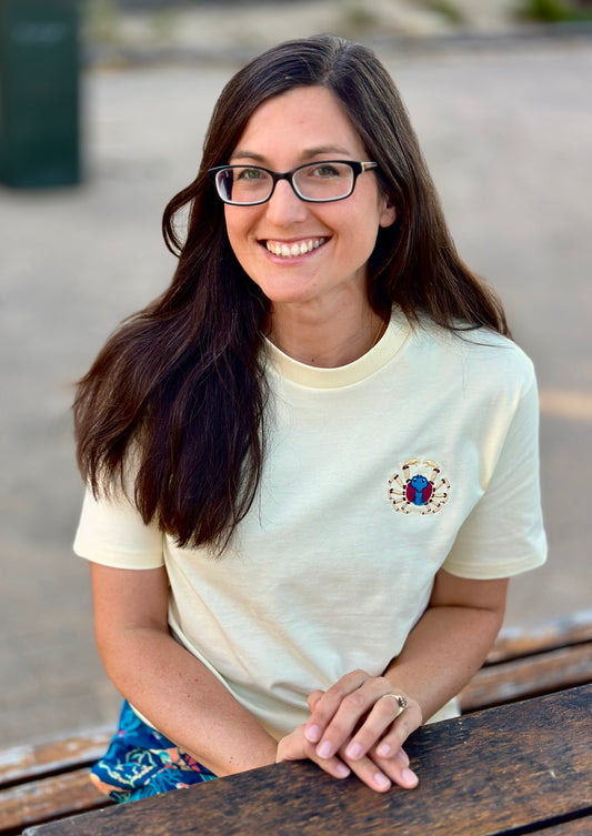 Woman wearing a white t-shirt with a embroidered soldier crab, sitting on a wooden bench outdoors.