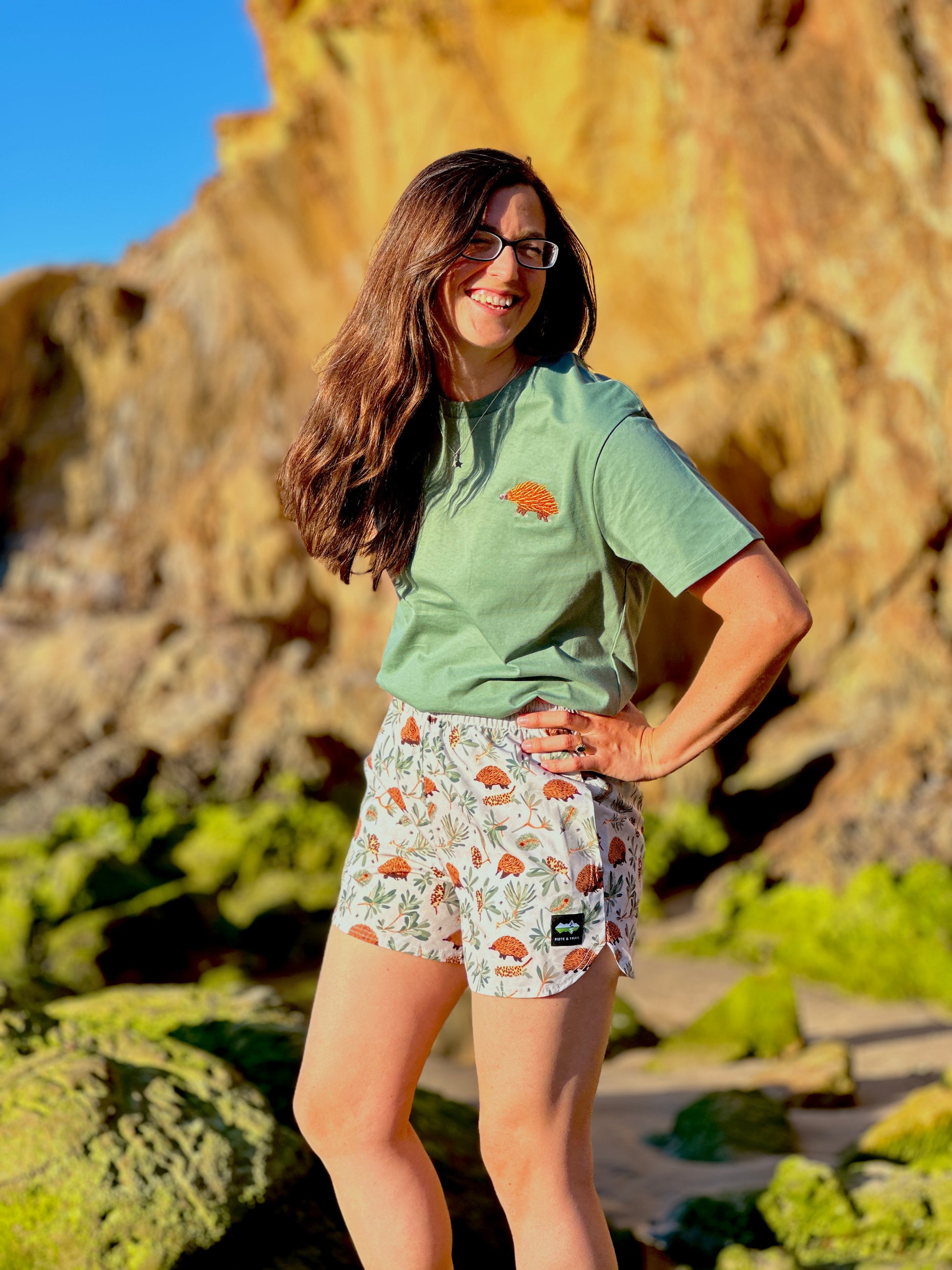 Woman standing in a natural setting wearing a green t-shirt and patterned shorts.
