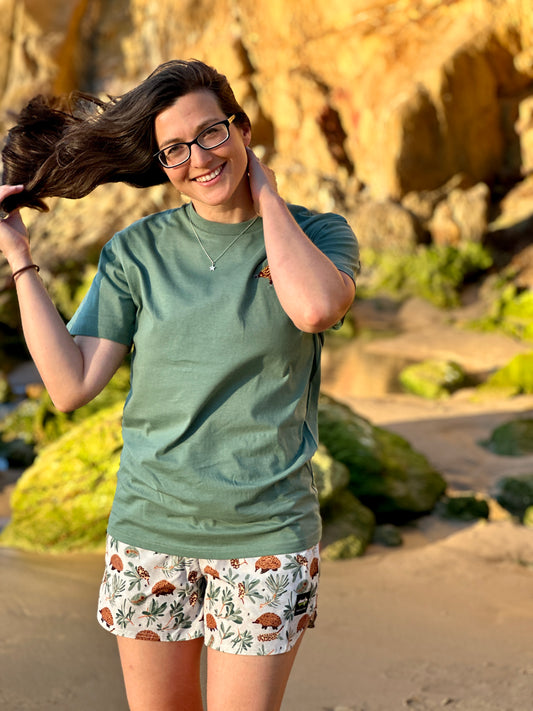 Woman on a beach wearing a green t-shirt and patterned shorts, with rocks and sand in the background.